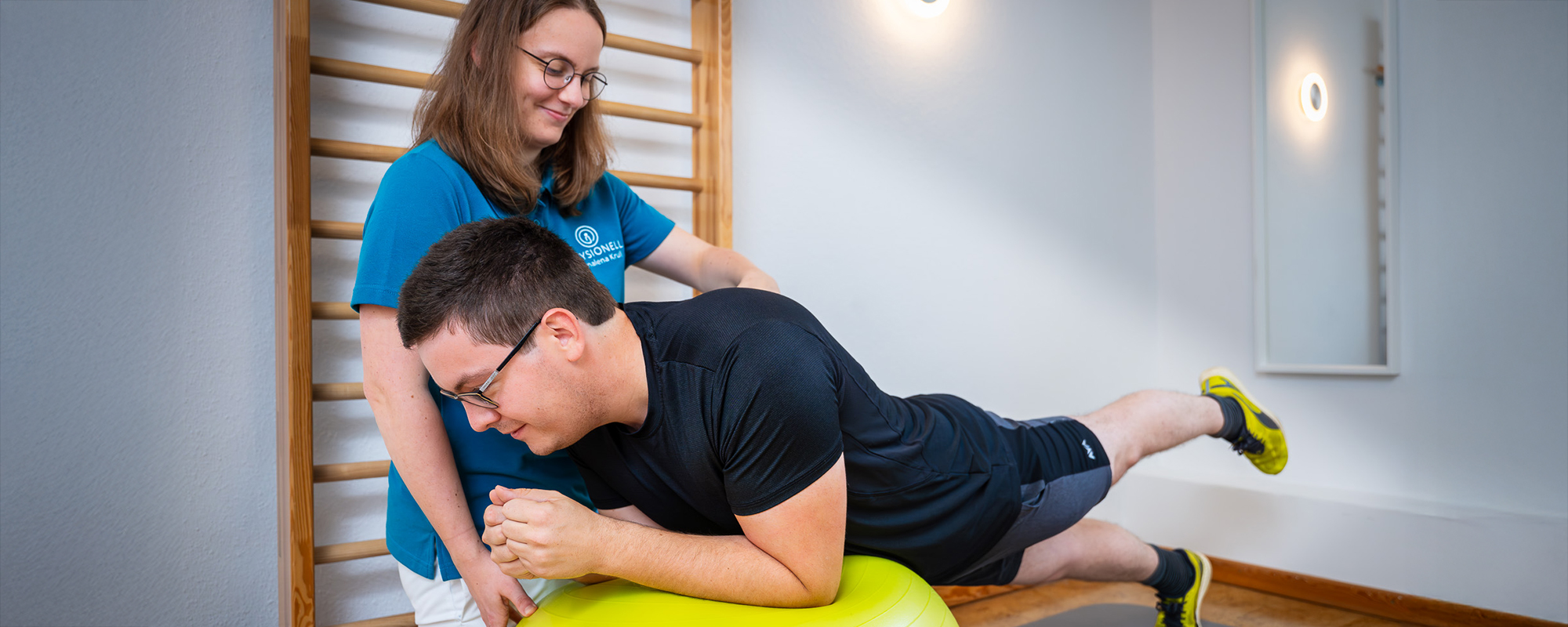 Allgemeine Krankengymnastik Physiotherapeutin Julia Neelsen steht bei einem Patienten während diese leicht gebeugt eine Übung der Krankengymnastik macht.